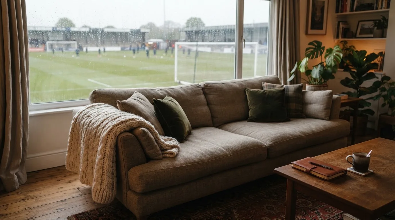 Ein Sofa mit Wolldecke steht vor einem grossen Fenster, draussen Regen und in der Ferne ein Fussballplatz.