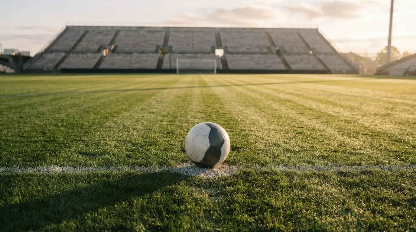 Ein Fussball liegt auf dem Anstosspunkt eines leeren Rasenplatzes im frühen Morgenlicht, dahinter erhebt sich eine menschenleere Tribüne.