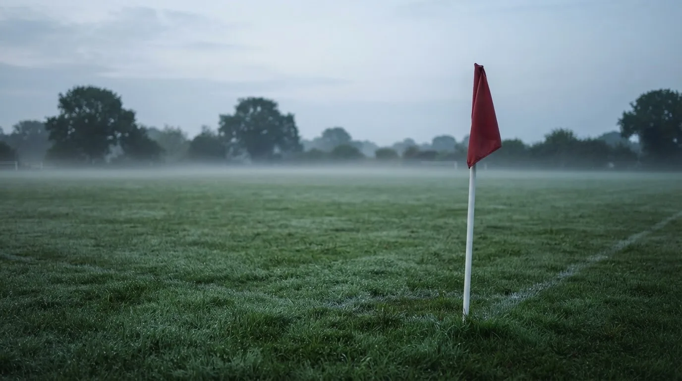 Eine Eckfahne steht am Morgen auf nassem Rasen, Tautropfen hängen im Gras und über dem Platz liegt leichter Dunst.