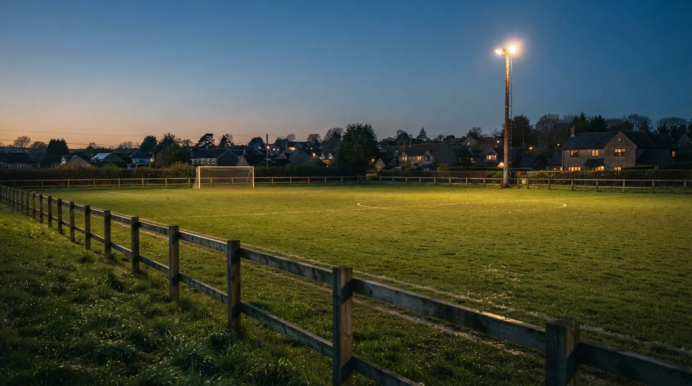 Ein kleiner Dorf-Fussballplatz liegt in der Dämmerung unter einem hohen Flutlichtmast, am Spielfeldrand steht ein Holzzaun.