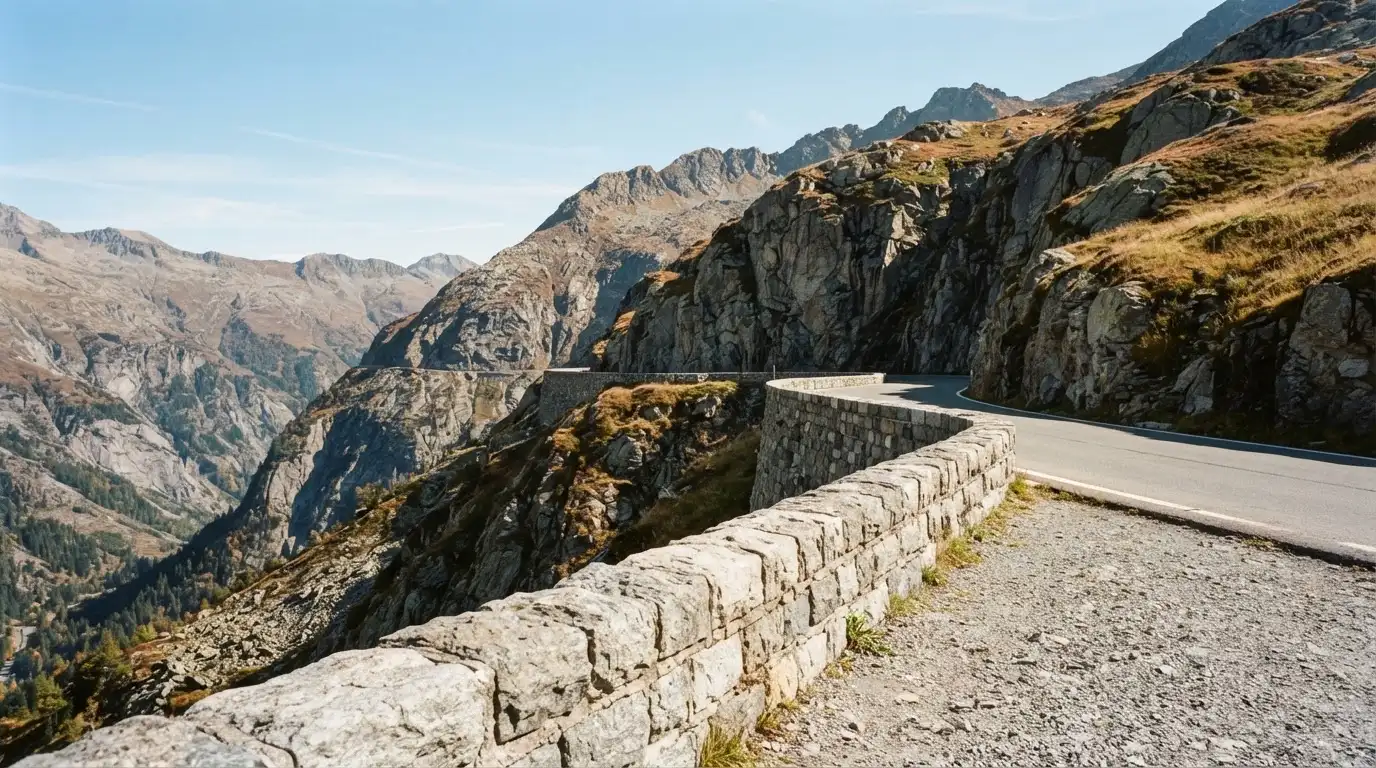 Eine Passstrasse im Hochgebirge säumt eine niedrige Steinmauer, dahinter ragen Felswände auf und der Herbsthimmel ist klar.