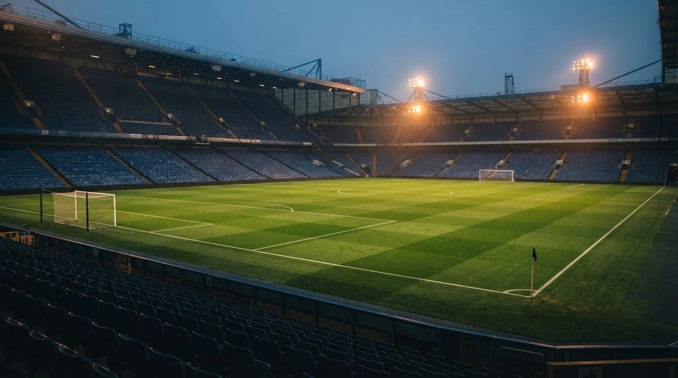 Leere Tribünenränge eines Fussballstadions in der Abenddämmerung, über dem Rasen leuchten die Flutlichter.