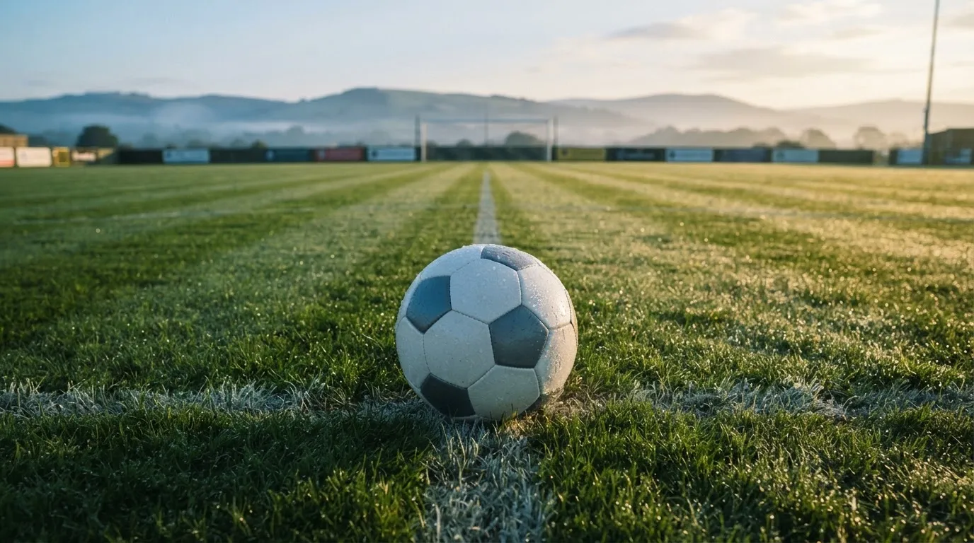 Ein Fussball liegt am frühen Morgen auf dem Anstosspunkt eines taunassen Rasens, im Hintergrund erheben sich Hügel im Dunst.