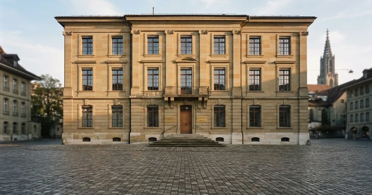 Die Fassade eines Sandsteingebäudes mit hohen Fenstern im weichen Licht des Vormittags, davor ein leerer Platz.