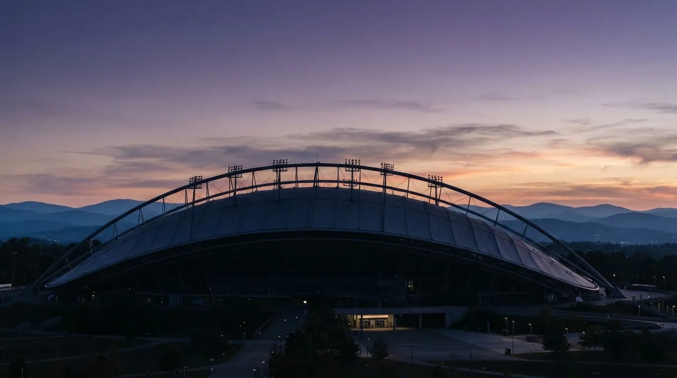 Die Silhouette eines Stadiondachs vor dem violetten Abendhimmel, dahinter eine Hügelkette.