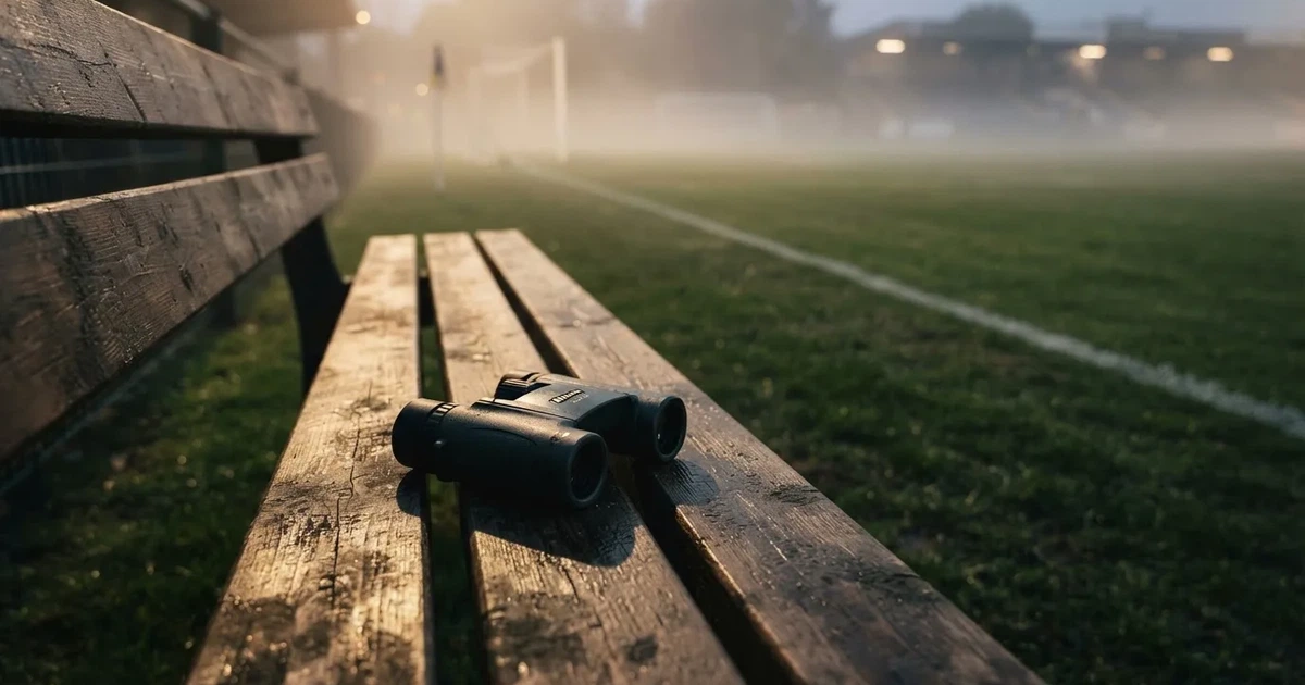 Ein Fernglas liegt auf einer leeren Holzbank an der Seitenlinie eines Fussballfelds, vom Rasen steigt Nebel auf.