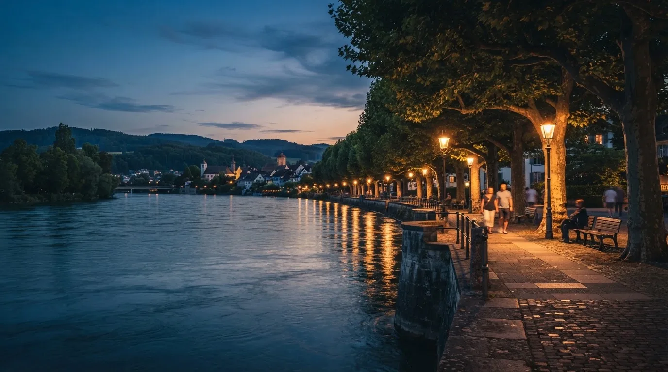 Eine Rheinpromenade mit hohen Bäumen am Abend, das Wasser spiegelt das Licht der ersten Laternen.