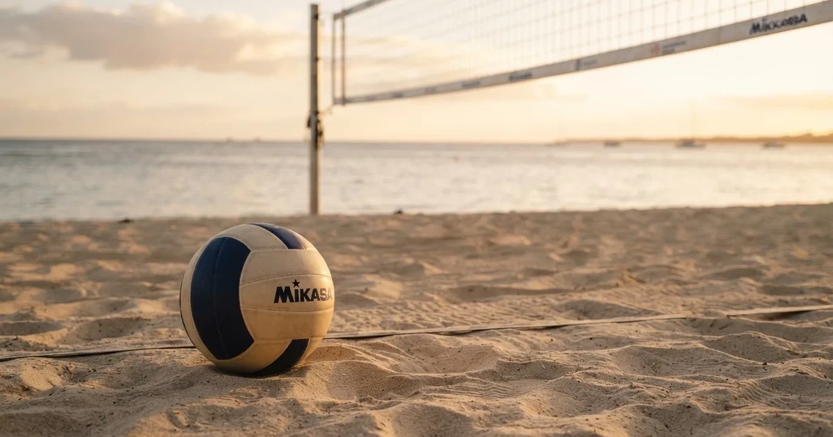 Ein Volleyball liegt auf feinem hellem Sand, ein gespanntes Netz verschwimmt dahinter, das Licht kommt von einem weiten Meer.