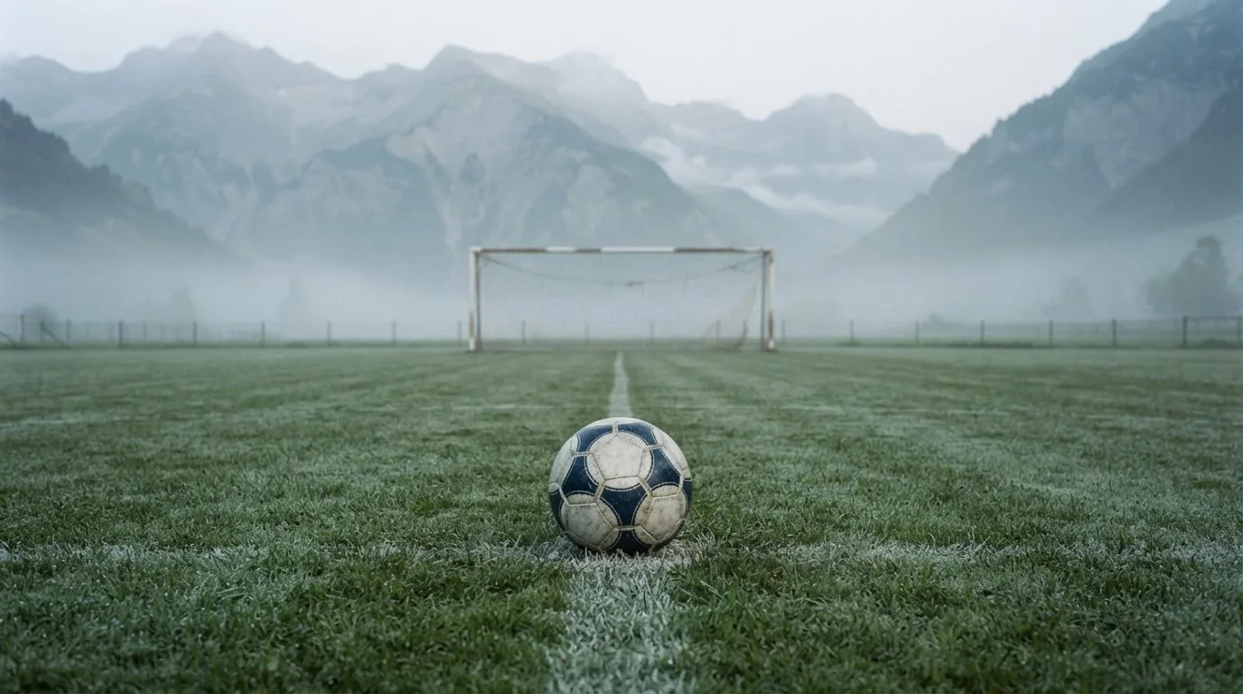 Ein einzelner Fussball auf dem Mittelpunkt eines leeren Rasens im Morgennebel, hinter dem Tor ragen Berge auf