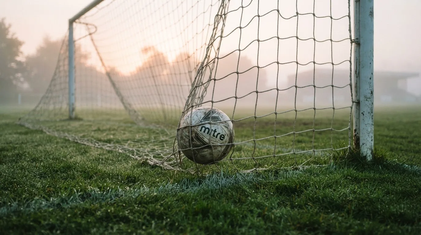 Ein Fussball liegt im Netz hinter einem Tor, weicher Morgennebel hängt über dem Rasen