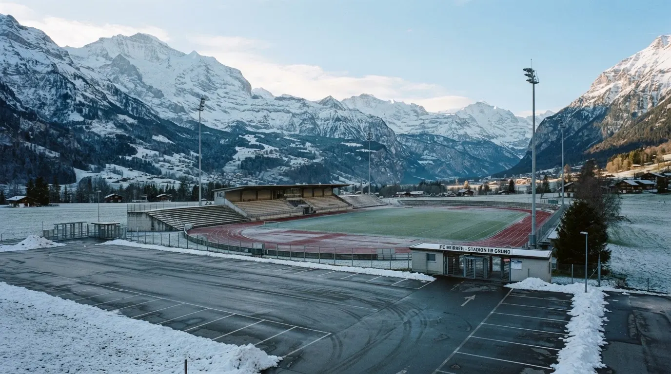 Schneebedeckte Berge ragen hinter einem kleinen Stadion in einem Alpental auf, im Vordergrund ein leerer Parkplatz am Morgen.