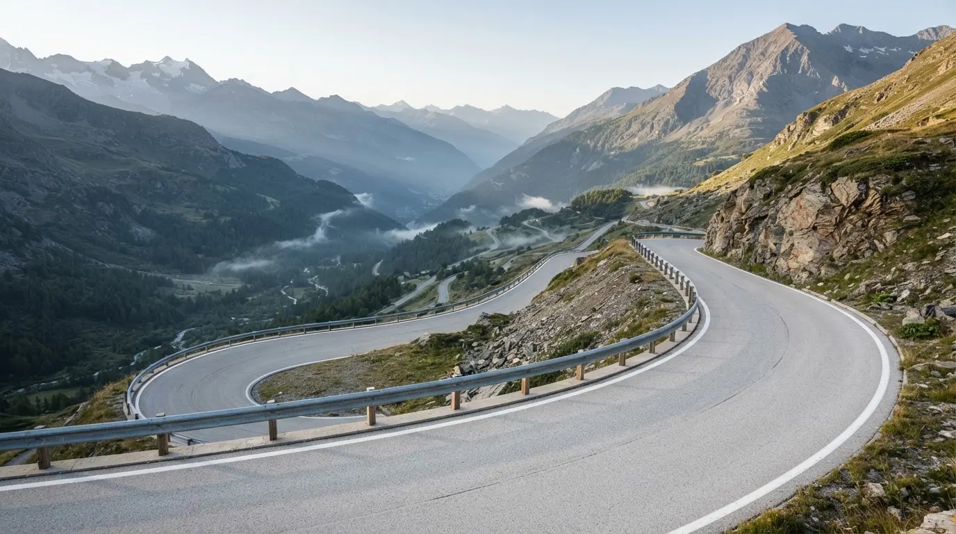Eine Passstrasse in den Alpen mit enger Kurve und Leitplanke, darüber ein klarer Morgenhimmel und einzelne Nebelschwaden im Tal.
