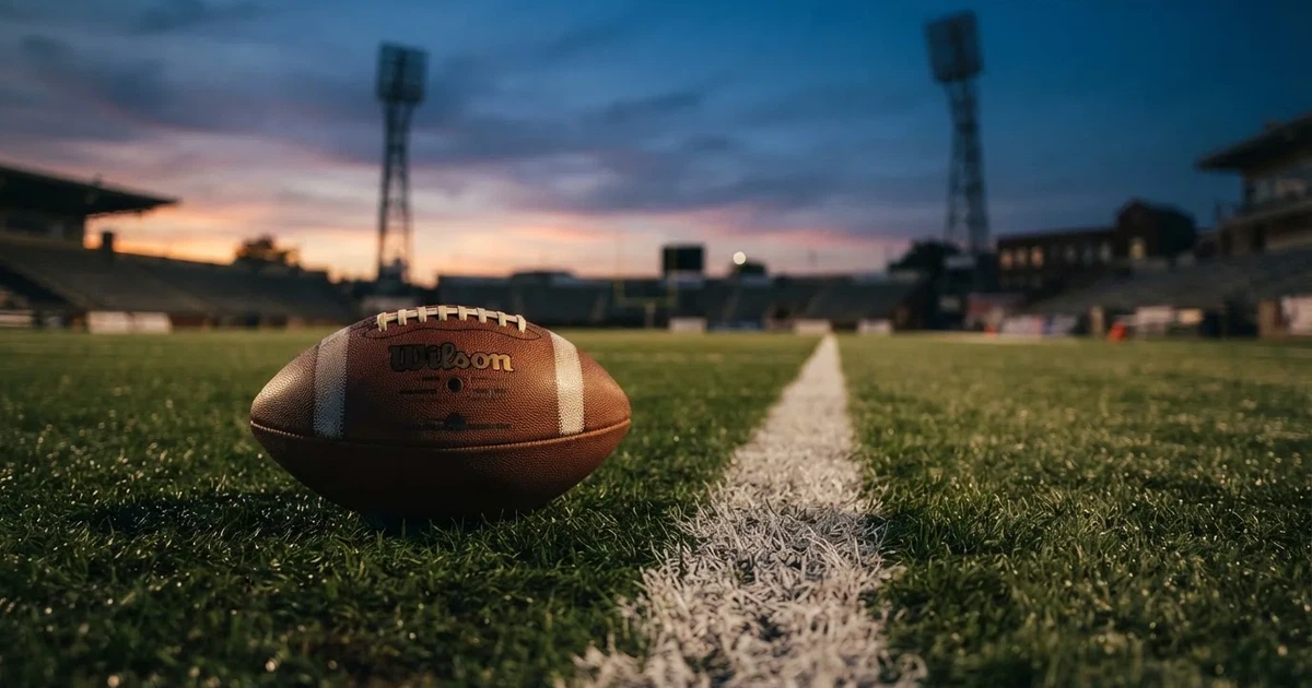 Ein ovaler Football liegt auf frisch gemähtem Rasen neben einer weissen Linie, im Hintergrund stehen leere Flutlichtmasten im Abendlicht.