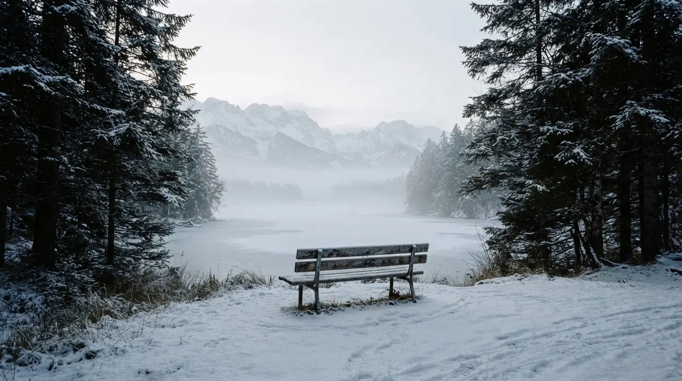 Ein zugefrorener Bergsee im Morgendunst, am Ufer steht eine einsame Holzbank zwischen verschneiten Tannen.