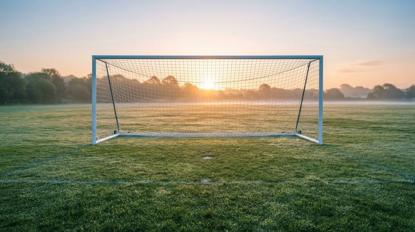 Ein Fussballtor mit Netz in der Totalen auf einem leeren Platz, Tau glänzt im Gras und die Morgensonne steht tief.