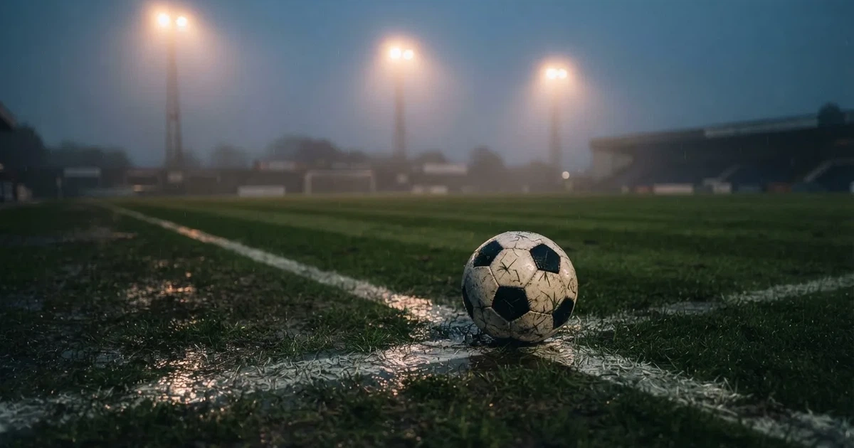 Ein Fussball liegt auf der Strafraumlinie eines nassen Rasens, dahinter stehen Flutlichtmasten im aufsteigenden Nebel.