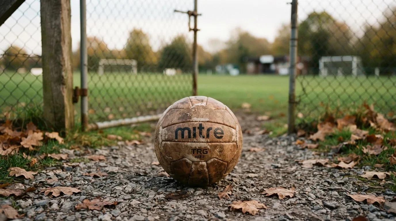 Ein abgenutzter Fussball liegt auf einem Kiesweg neben dem Eingang eines Vereinsplatzes, Laub liegt auf dem Boden.