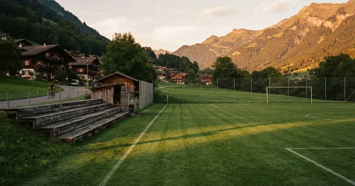 Ein kleiner Fussballplatz mit einfacher Holztribüne und Rasenstreifen am Rand einer Ortschaft, im Hintergrund ein bewaldeter Hügel im Abendlicht.