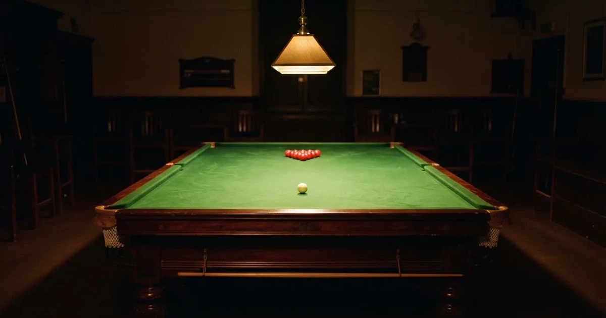 Ein Snookertisch mit grünem Tuch und einem Dreieck aus roten Kugeln unter einer tief hängenden Lampe in einer leeren Halle.