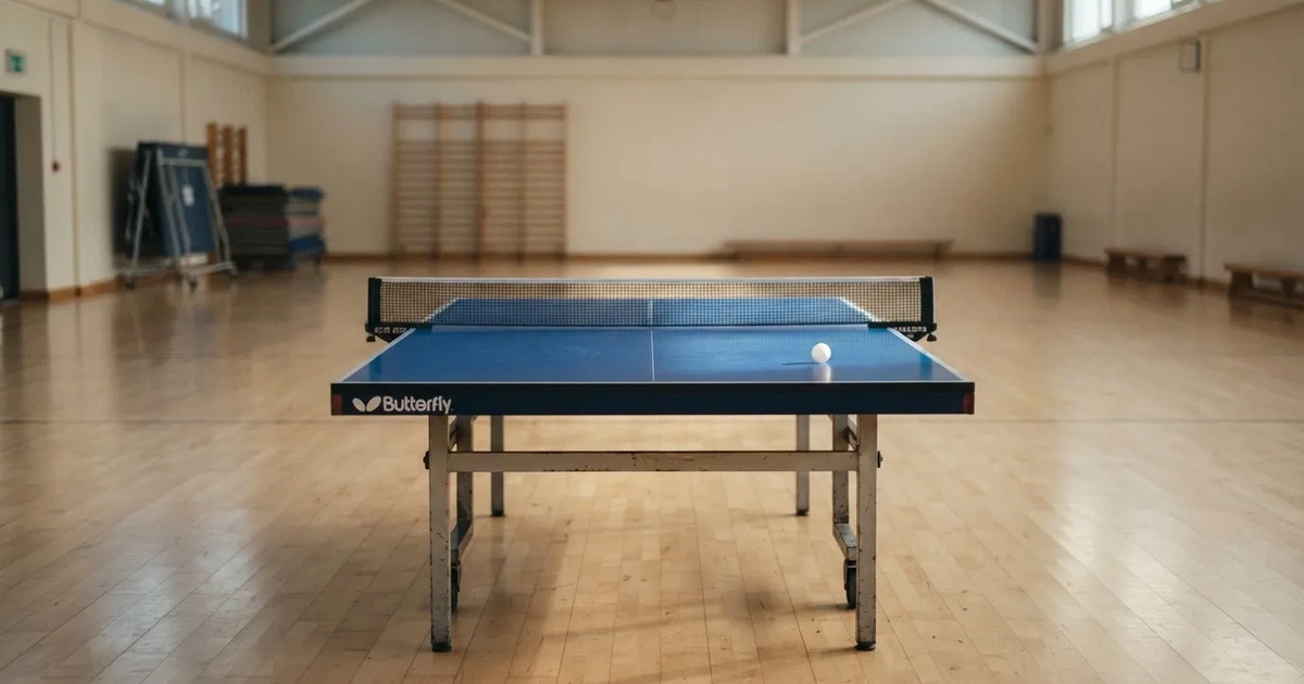 Eine Tischtennisplatte mit Netz in einer leeren Sporthalle, ein heller Ball liegt auf einer Seite, Deckenlicht spiegelt sich im Hallenboden.