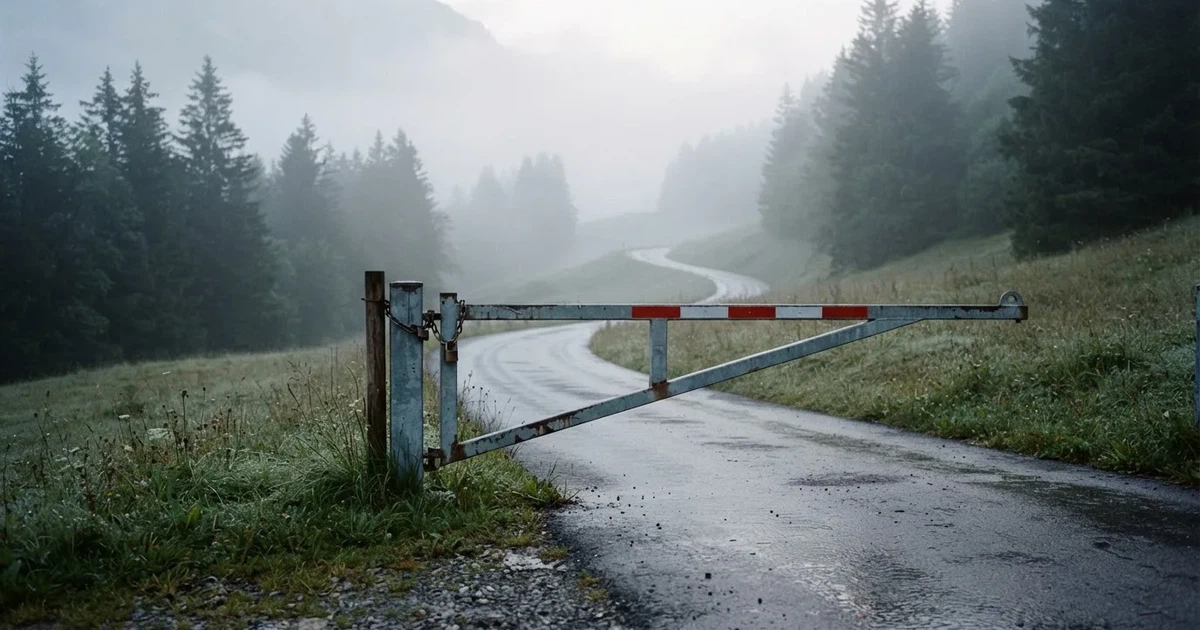 Eine geschlossene Schranke steht an einer ruhigen Bergstrasse im Morgennebel, dahinter Tannen und eine nasse Wiese.