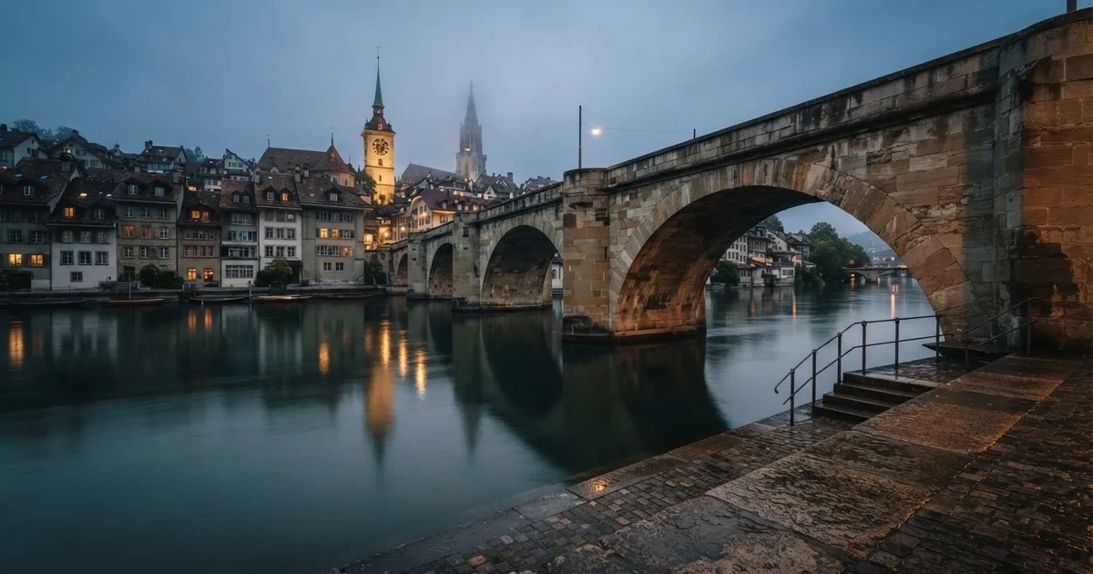 Eine alte Steinbrücke führt über einen Fluss in einer Schweizer Stadt, das Wasser ist ruhig und das Licht kühl.