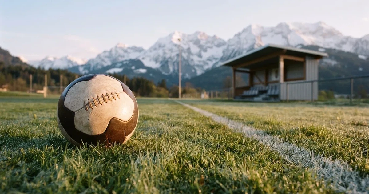 Ein Fussball liegt am frühen Morgen auf dem Rasen eines leeren Fussballfeldes, dahinter eine kleine Tribüne und schneebedeckte Berge.