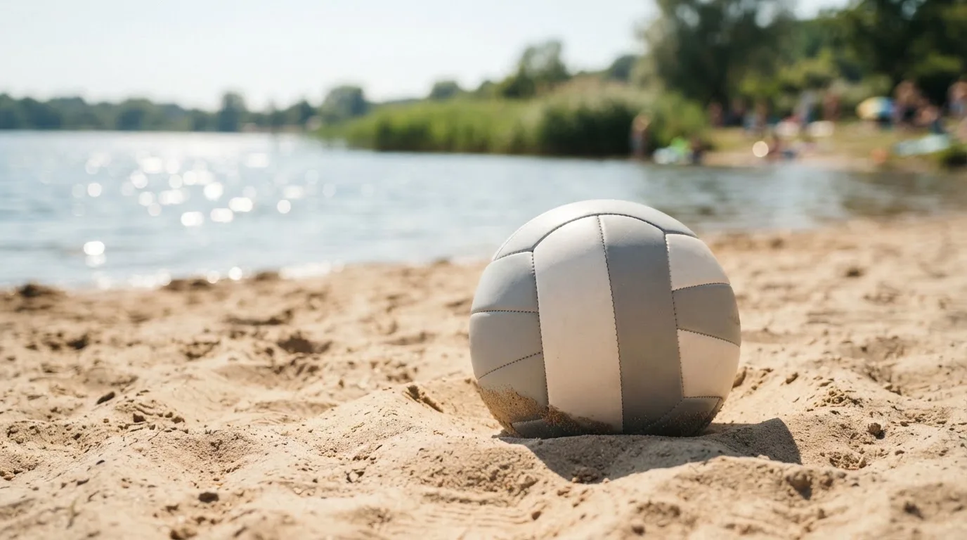 Ein Volleyball liegt auf dem Sand eines Strandfeldes am Seeufer, im Hintergrund glitzert das Wasser.