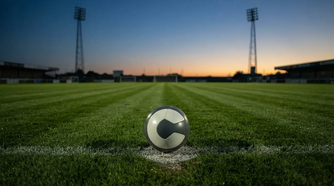 Ein Fussball liegt auf dem Mittelpunkt eines leeren Rasenplatzes, im Hintergrund ragen Flutlichtmasten in die Abenddämmerung