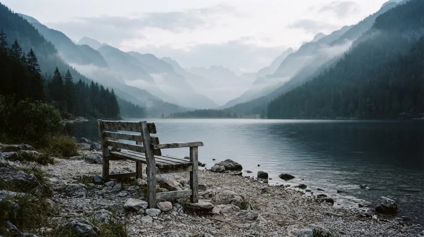 Eine Holzbank am Ufer eines ruhigen Bergsees, am gegenüberliegenden Ufer steigen dunkle Hänge in den Frühnebel