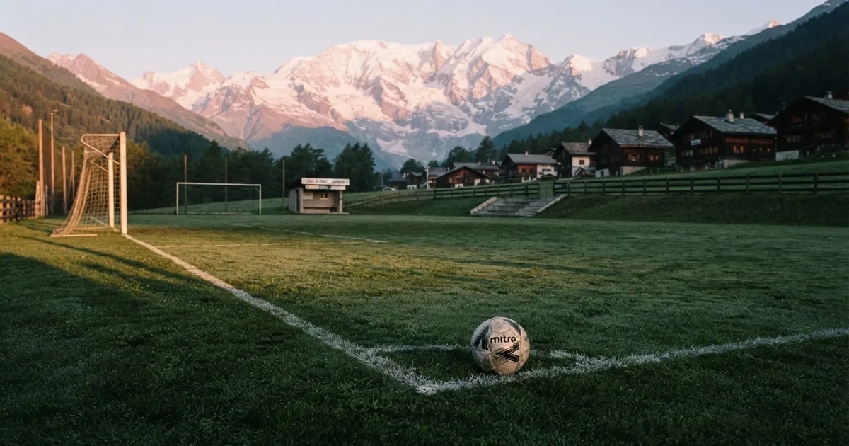 Ein Dorf-Sportplatz mit einem Ball am Strafraumrand, dahinter schneebedeckte Alpengipfel im klaren Morgenlicht