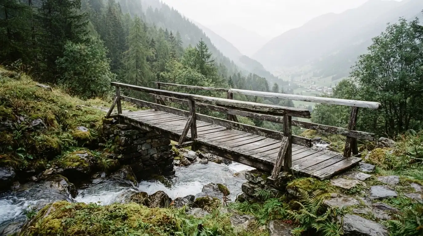 Eine alte Holzbrücke über einen Gebirgsbach, Moos auf den Steinen, Nebelschwaden hängen im Tal