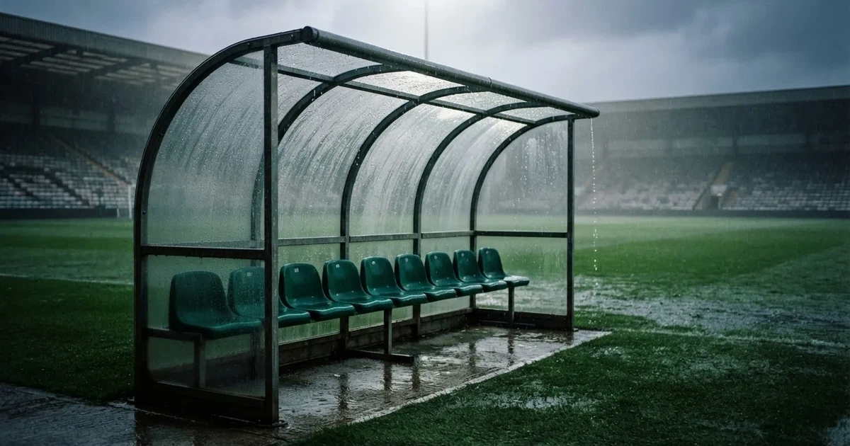 Eine leere Ersatzbank unter einem Plexiglasdach am Spielfeldrand, Regentropfen auf der Scheibe, dahinter ein Rasenplatz