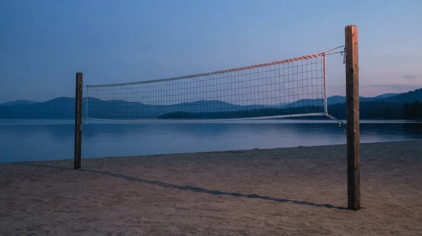 Ein Volleyballnetz steht an einem Seeufer in der Abenddämmerung, der Sand ist leer und das Wasser glatt.