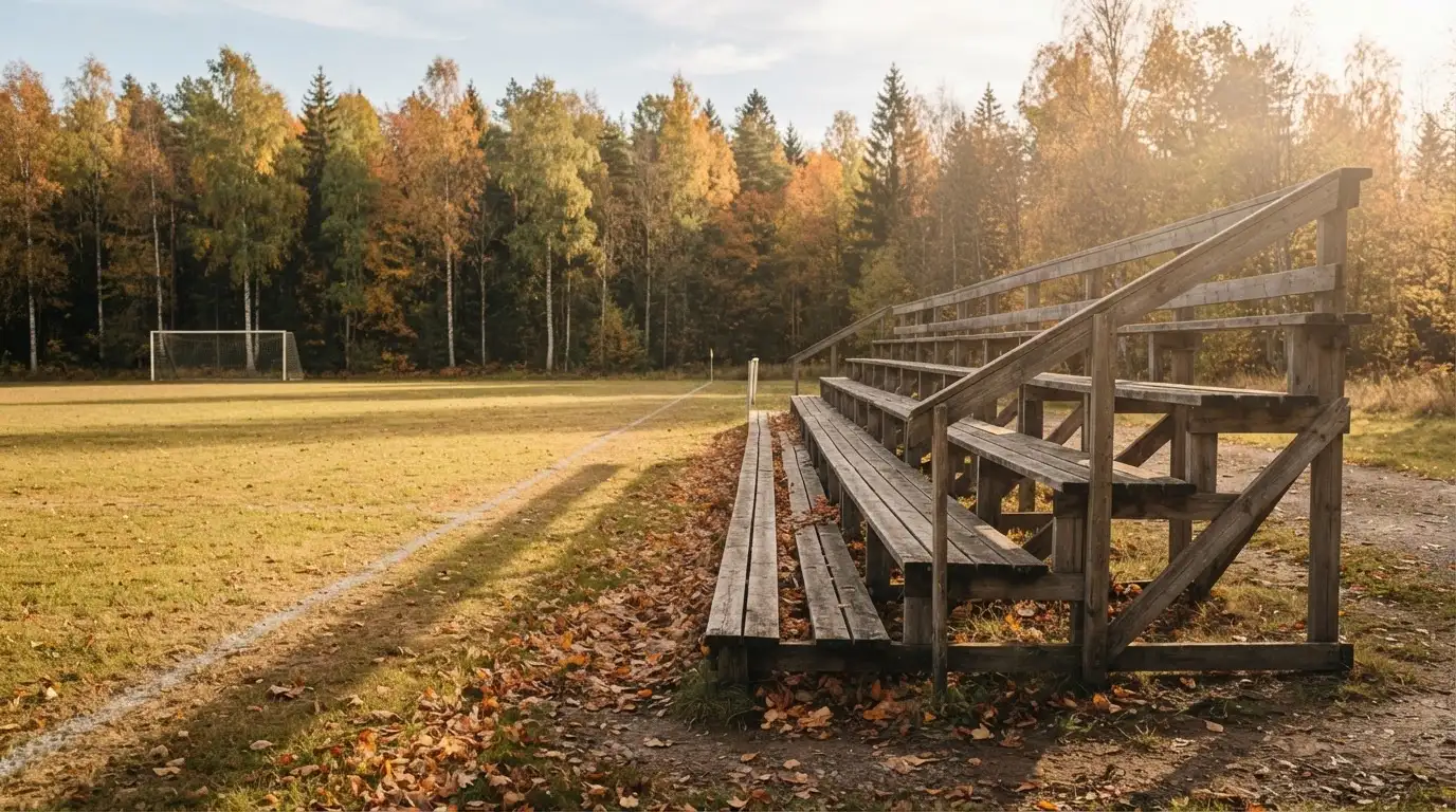 Eine leere Holztribüne steht am Waldrand neben einem kleinen Sportplatz im Herbstlicht.