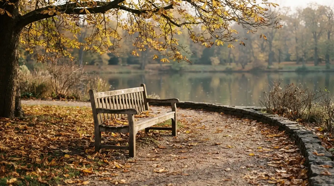 Eine Parkbank unter einer Kastanie, Herbstlaub bedeckt den Kiesweg, im Hintergrund ein stiller Teich.