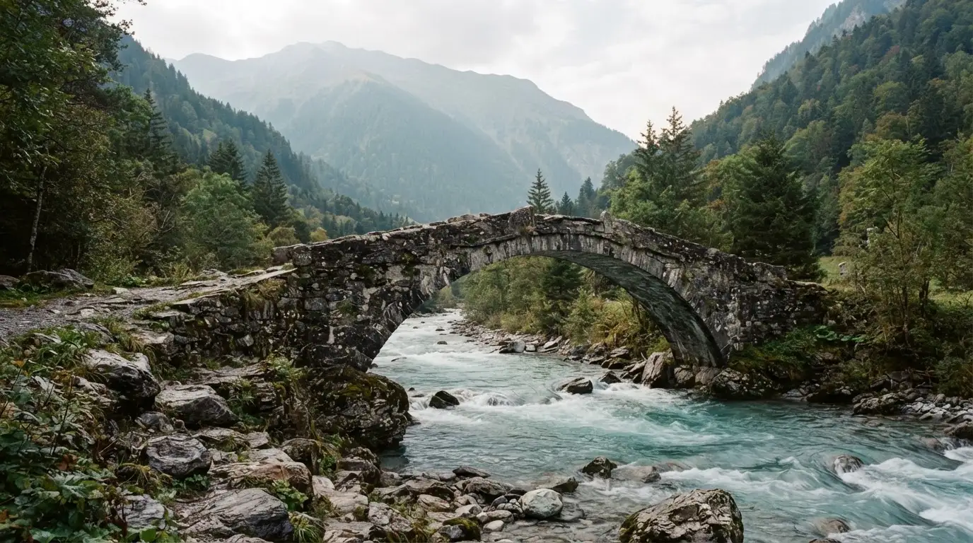Eine alte Steinbrücke überspannt einen Gebirgsfluss, im Hintergrund steigen bewaldete Hänge an.